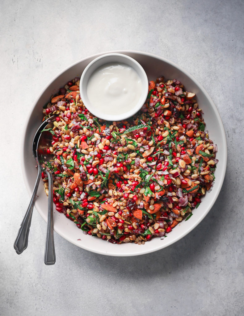 Grey bowl of Puy Lentil, Pomegranate and kamut grain salad, with a pot of yogurt and two spoons