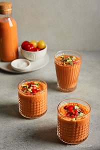 3 glasses of Spanish gazpacho soup on a beige background, bowl of tomatoes in rear of photo