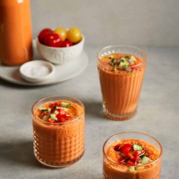 3 glasses of Spanish gazpacho soup on a beige background, bowl of tomatoes in rear of photo