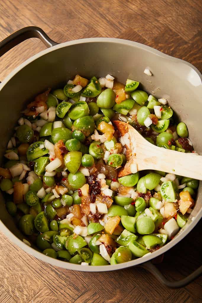 Image of diced green tomatoes and onions being added to part cooked chutney mixture in a large saucepan