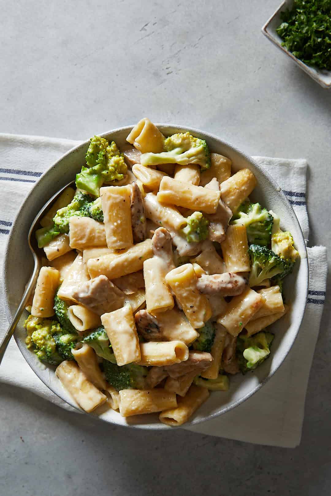Vegan Chicken & Broccoli Alfredo on a grey backdrop with a small bowl of parsely and a white striped linen