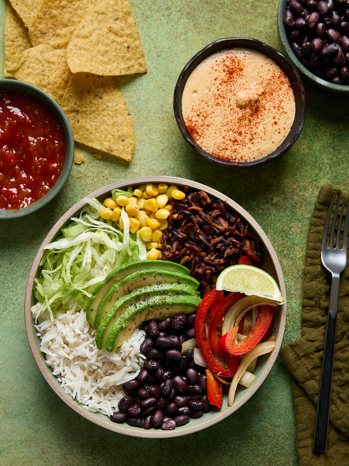 Vegan burrito bowl with white rice, black beans, avocado slices, shredded lettuce, corn, tofu beef crumbles, and garlic-sautéed peppers and onions, served with lime wedge, dairy-free queso, and a side of extra black beans