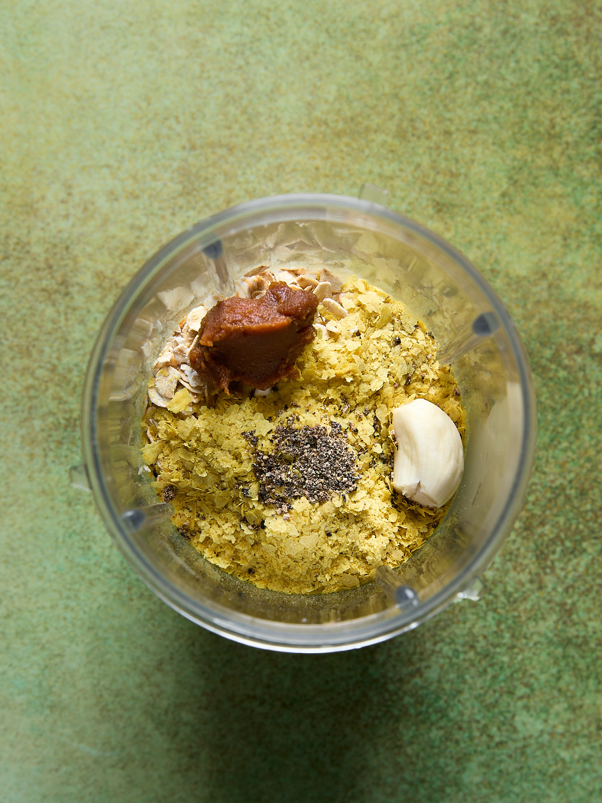 Blender cup filled with oats, nutritional yeast, garlic, miso paste, and black pepper.