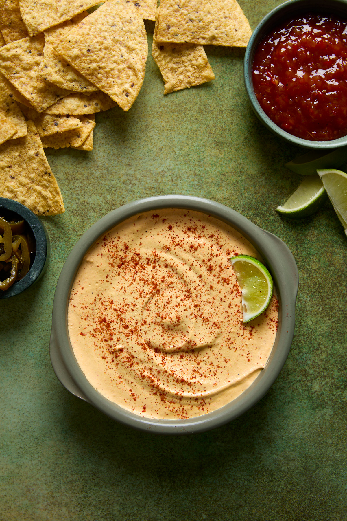 A bowl of creamy vegan nacho-style cashew queso topped with smoked paprika and a lime wedge, surrounded by tortilla chips, pickled jalapeños, salsa, and lime slices on a green surface.