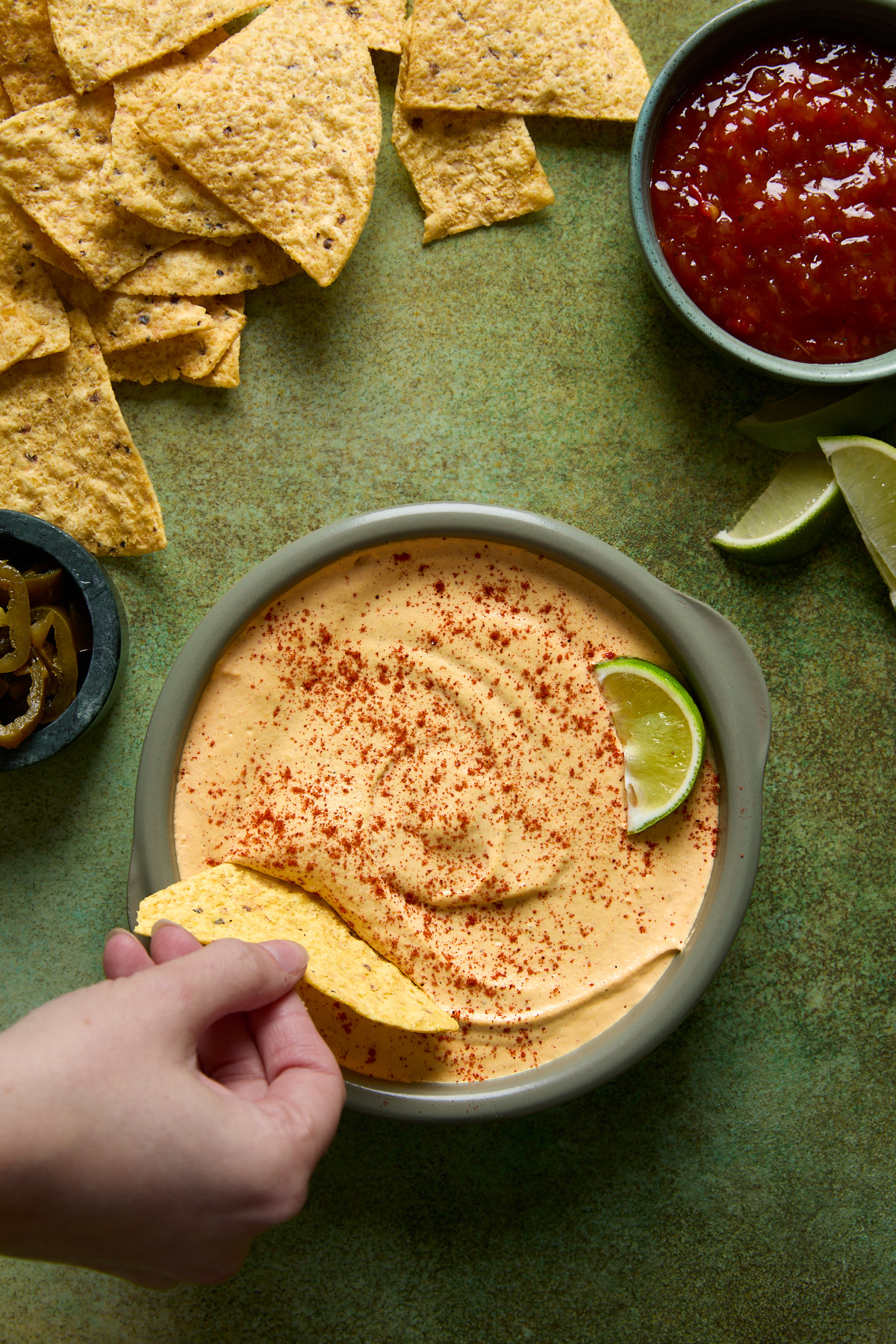 A bowl of creamy vegan nacho-style cashew queso topped with smoked paprika and a lime wedge, surrounded by tortilla chips, pickled jalapeños, salsa, and lime slices on a green surface.