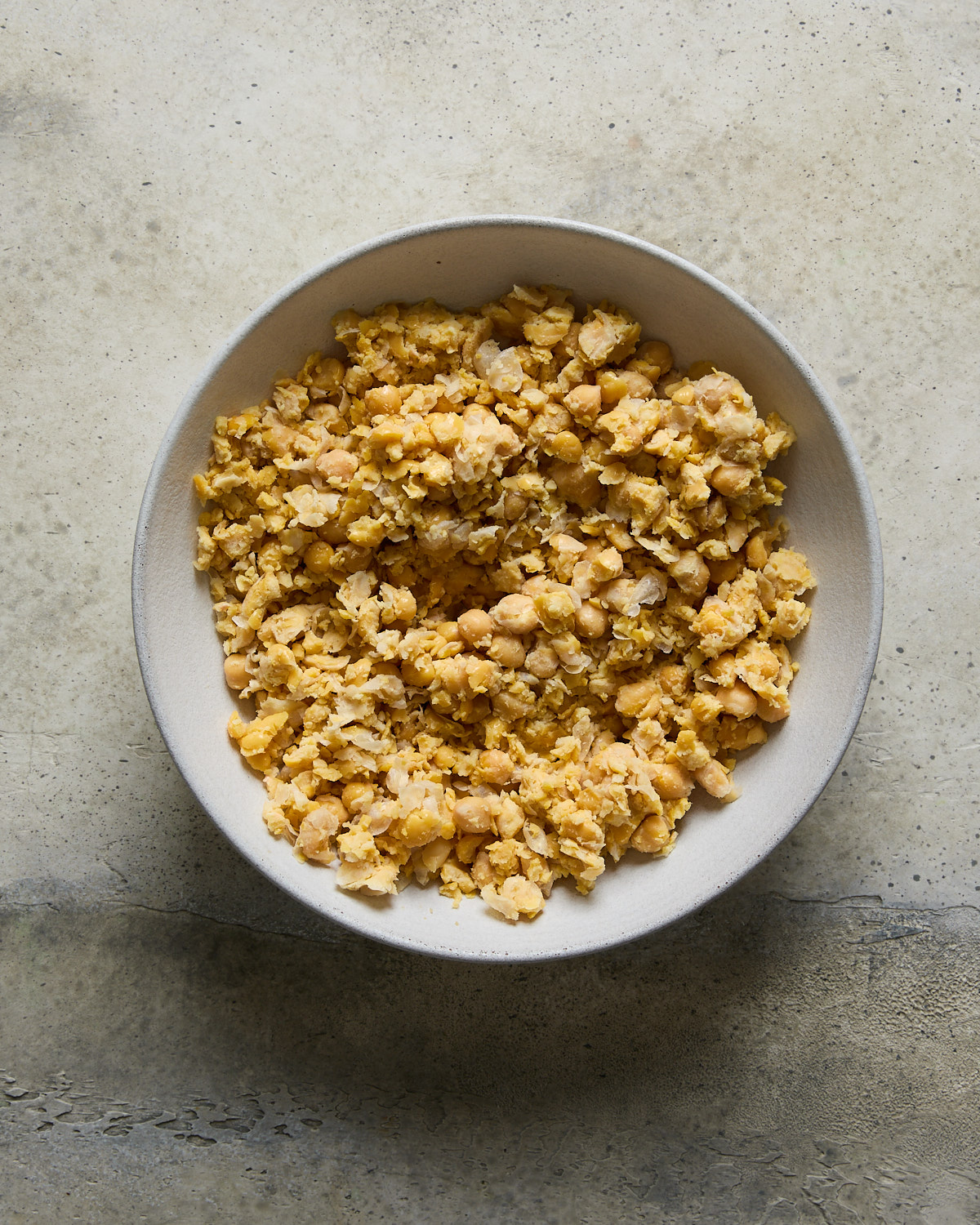 A white bowl filled with mashed chickpeas sits on a textured, light-colored surface. The chickpeas are roughly mashed.