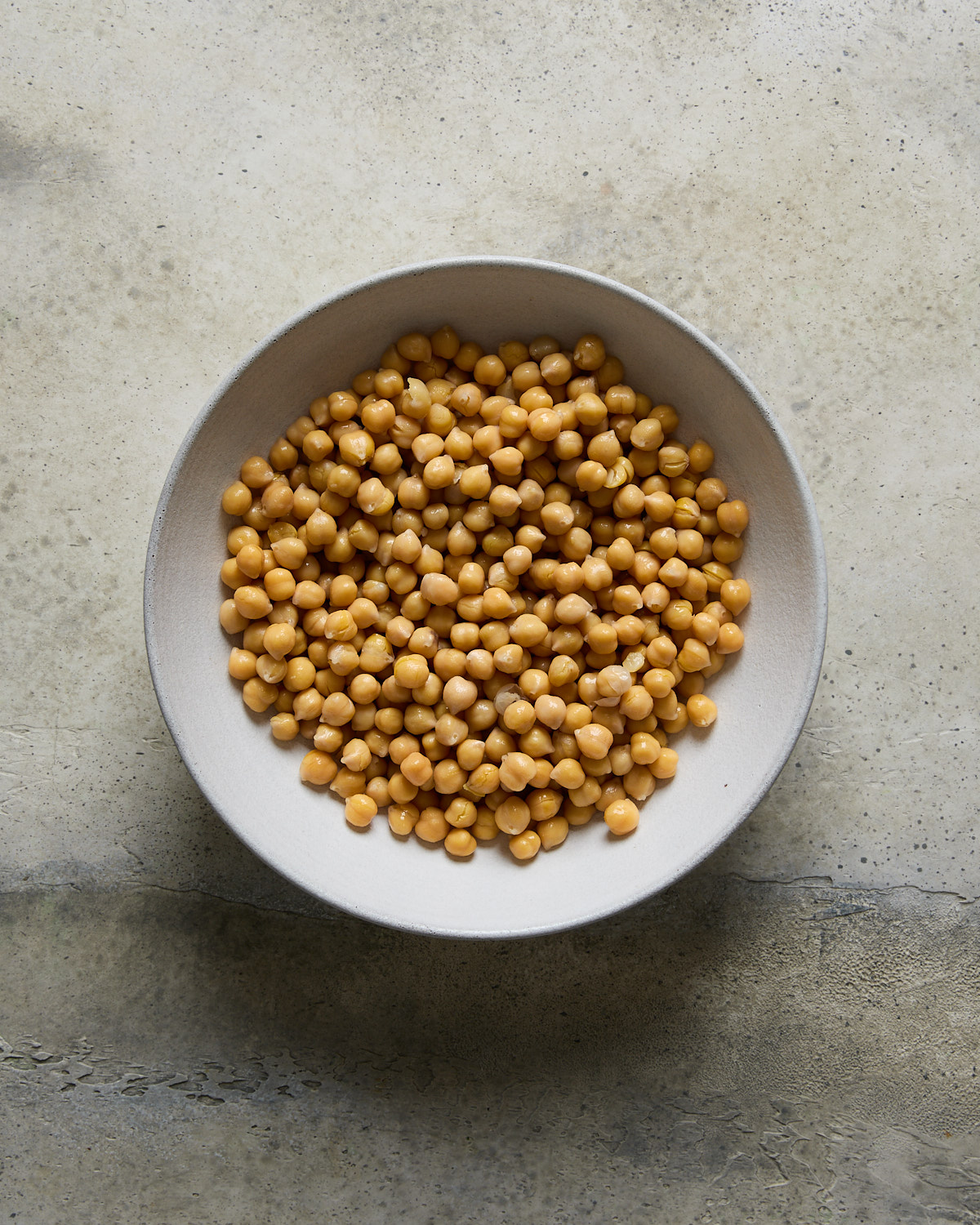 A white bowl filled with chickpeas is placed on a textured, light-colored surface.