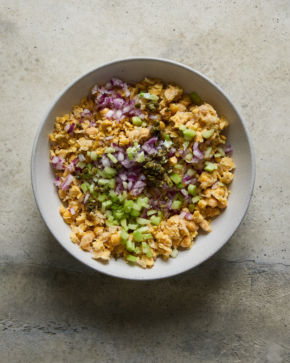 A bowl filled with mashed chickpeas, chopped red onions, diced celery, and a dollop of chopped capers, all in a light bowl.