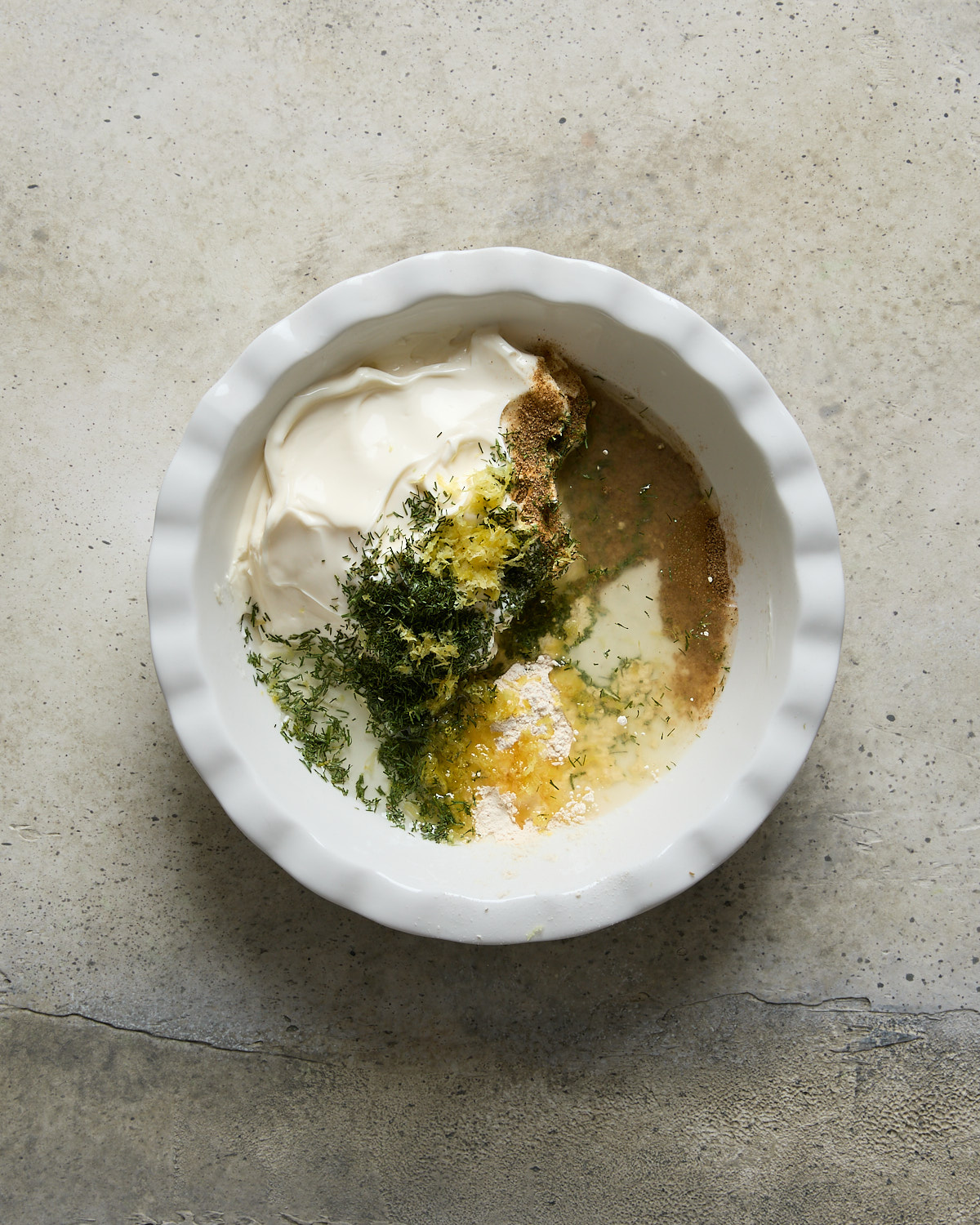 A white bowl on a concrete surface filled with separated ingredients: vegan mayo, herbs, spices, lemon zest, and lemon juice, ready to be mixed.