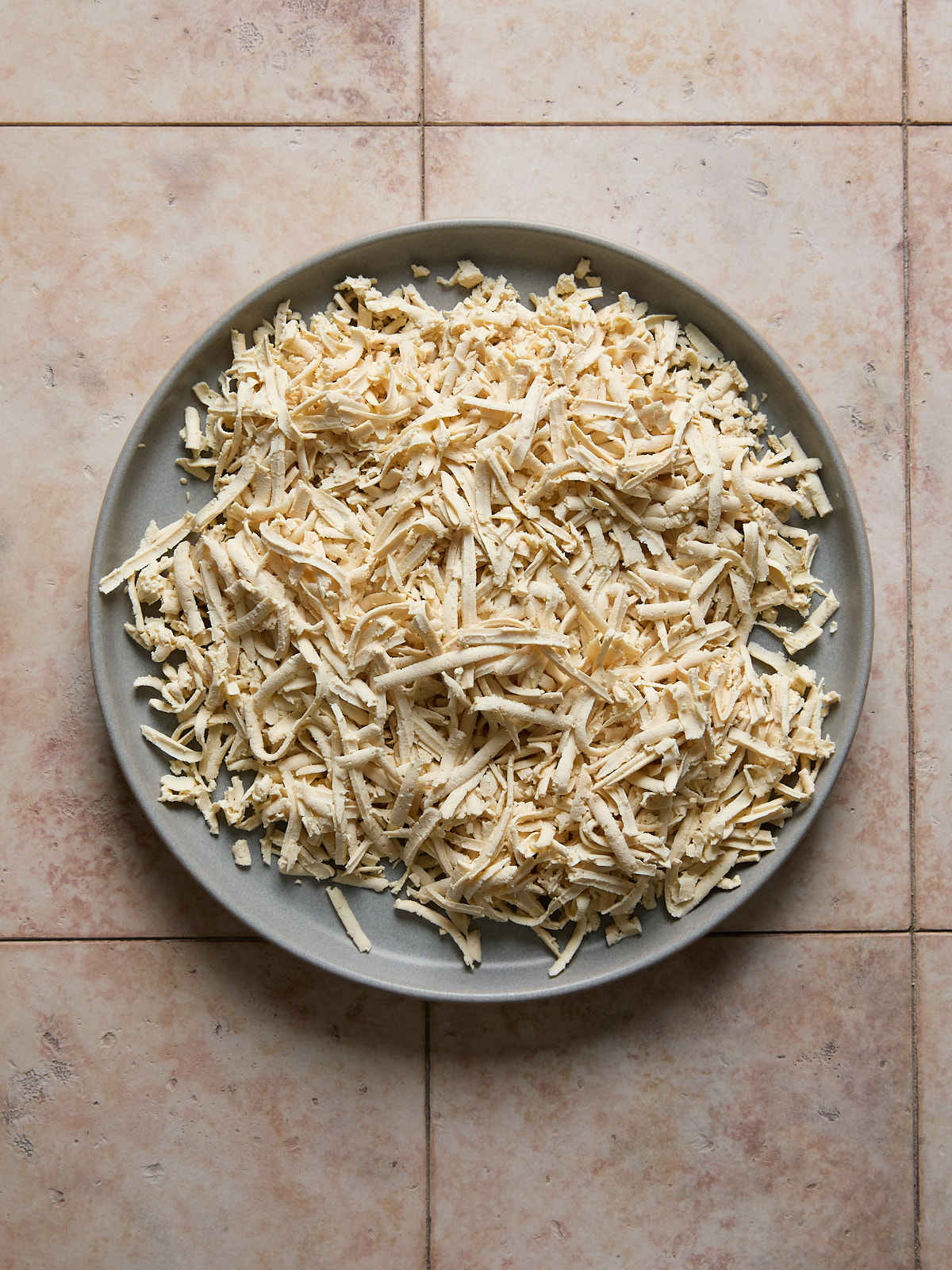 A gray plate filled with a large pile of shredded tofu sits on a lightly mottled beige tile surface.