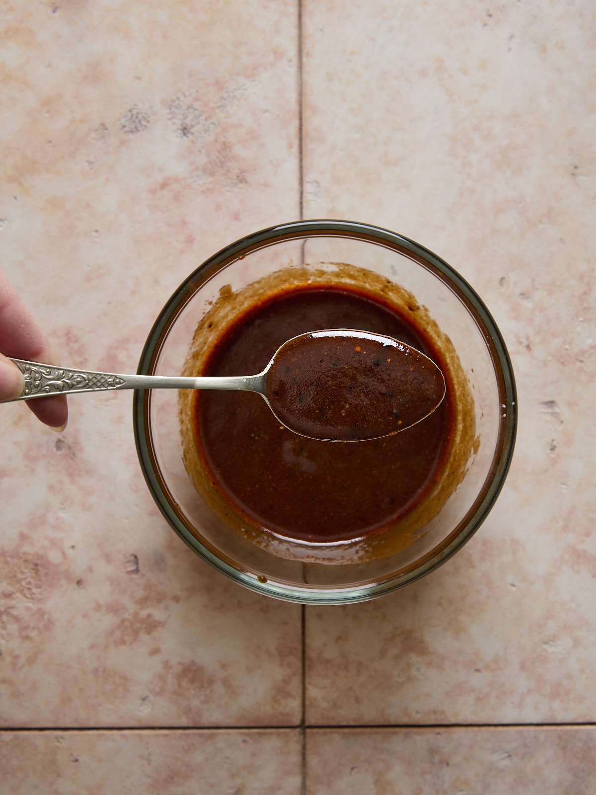 A hand holds a spoon above a glass bowl filled with the mixed tofu marinade, set on a light, marbled surface.