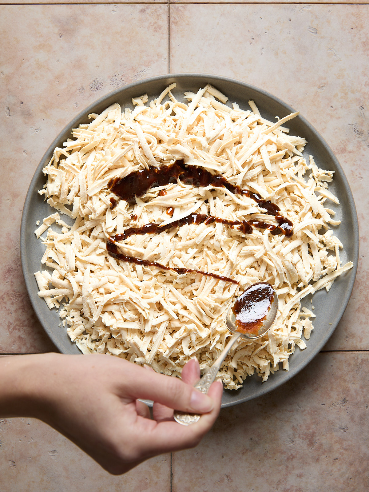 A hand sprinkles a spice from a spoon onto a plate of shredded white cheese, which has brown sauce drizzled on top, all placed on a tiled surface.