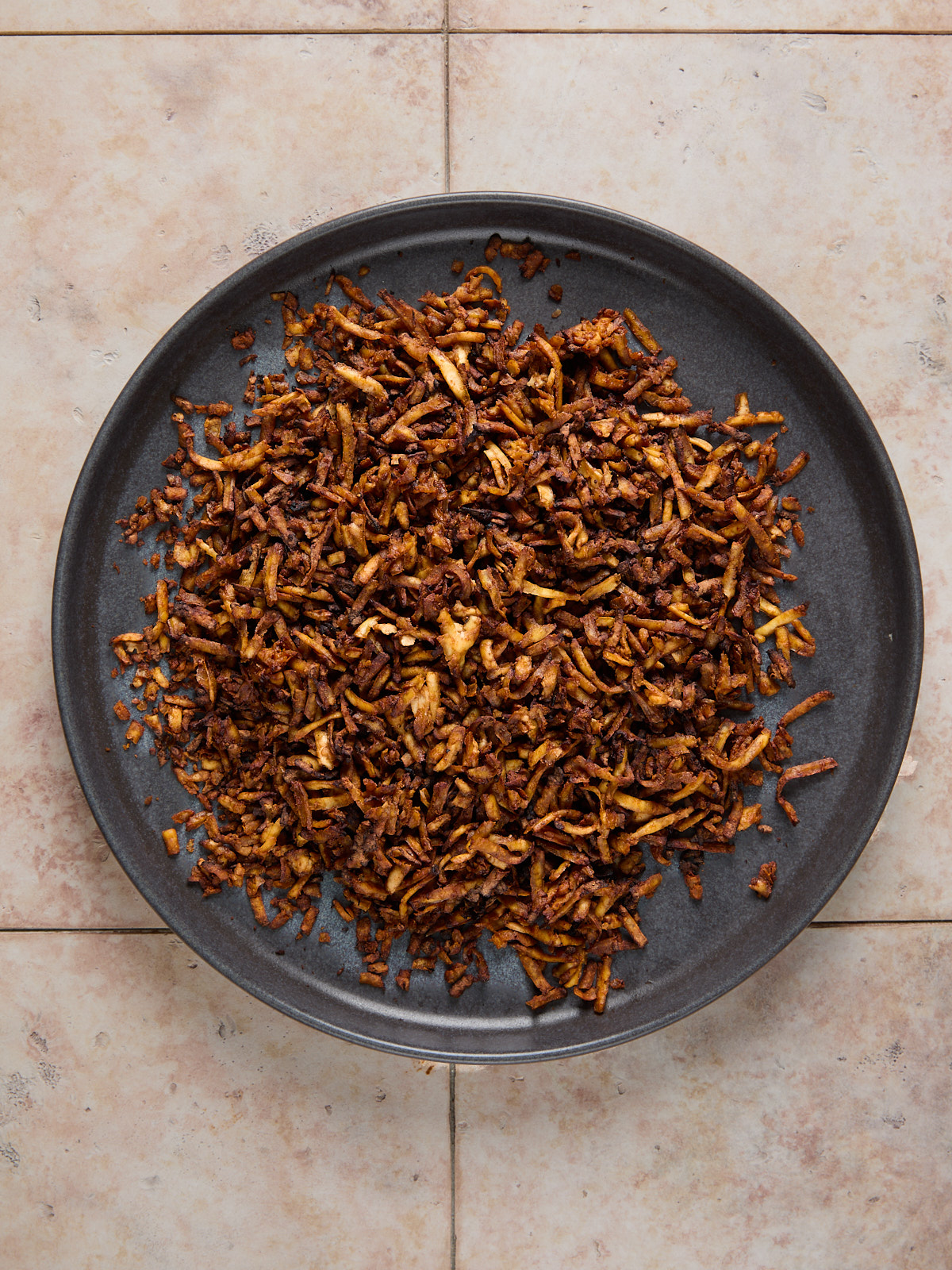 A dark plate filled with a heap of crispy, browned fried onions sits on a light-colored tiled surface.