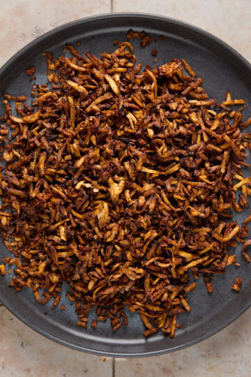 A dark plate filled with a heap of crispy, browned fried onions sits on a light-colored tiled surface.