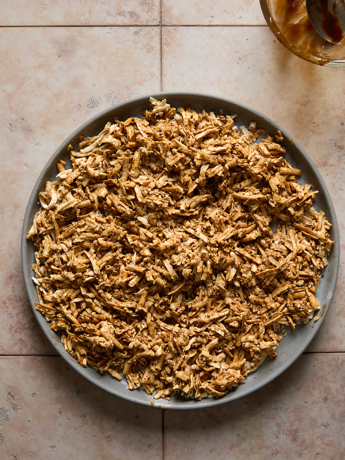 A grey plate filled with shredded, marinade coated shredded tofu sits on a tiled surface. A small portion of a glass bowl with sauce is visible in the upper right corner.