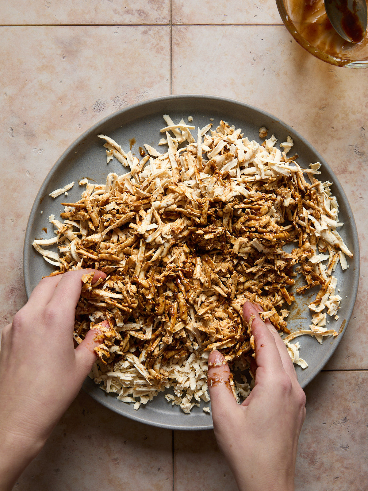 Two hands mix marinade into shredded tofu on a gray plate, sitting on a light pink tiled surface.