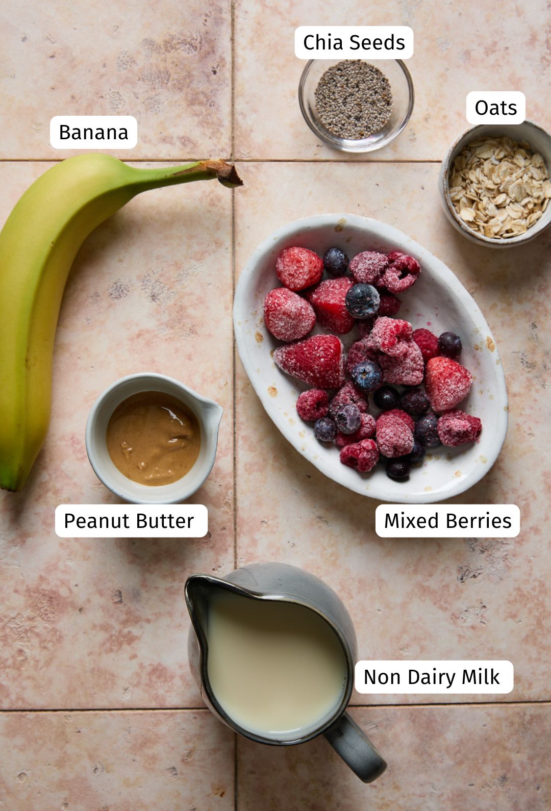 Overhead shot of smoothie ingredients including a ripe banana, frozen mixed berries, rolled oats, chia seeds, peanut butter, and non-dairy milk arranged on a tile background.