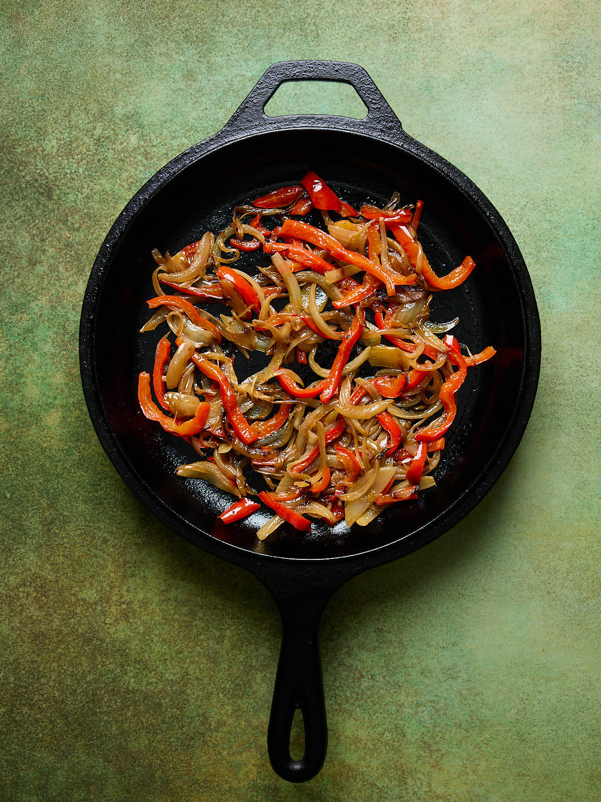 Cast iron skillet filled with sautéed red bell peppers and onions, glistening with olive oil on a green textured background.