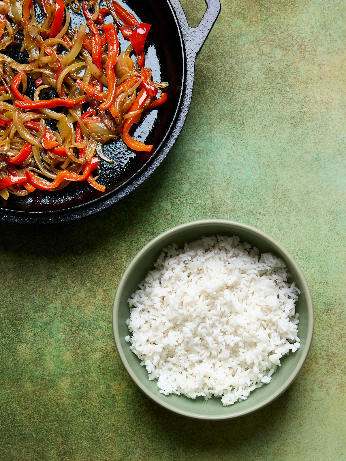 Cooked white rice in a green bowl next to a skillet of garlic-sautéed red peppers and onions, set on a green surface.