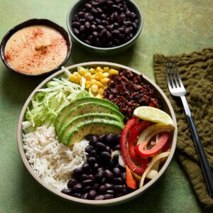 Vegan burrito bowl with white rice, black beans, avocado slices, shredded lettuce, corn, tofu beef crumbles, and garlic-sautéed peppers and onions, served with lime wedge, dairy-free queso, and a side of extra black beans.