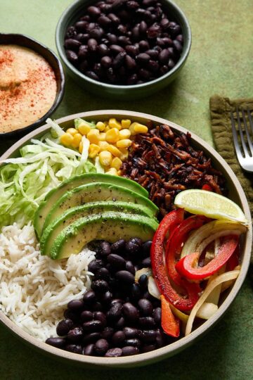 Vegan burrito bowl with white rice, black beans, avocado slices, shredded lettuce, corn, tofu beef crumbles, and garlic-sautéed peppers and onions, served with lime wedge, dairy-free queso, and a side of extra black beans.