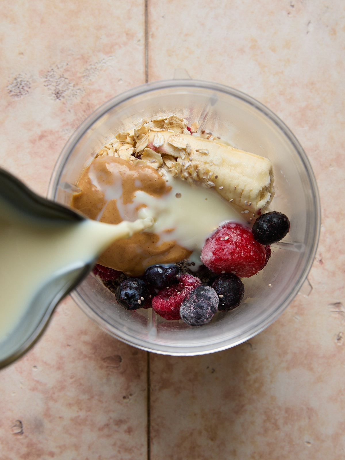 Close-up of banana, frozen berries, oats, chia seeds, and peanut butter in a blender cup with non-dairy milk being poured over the top.