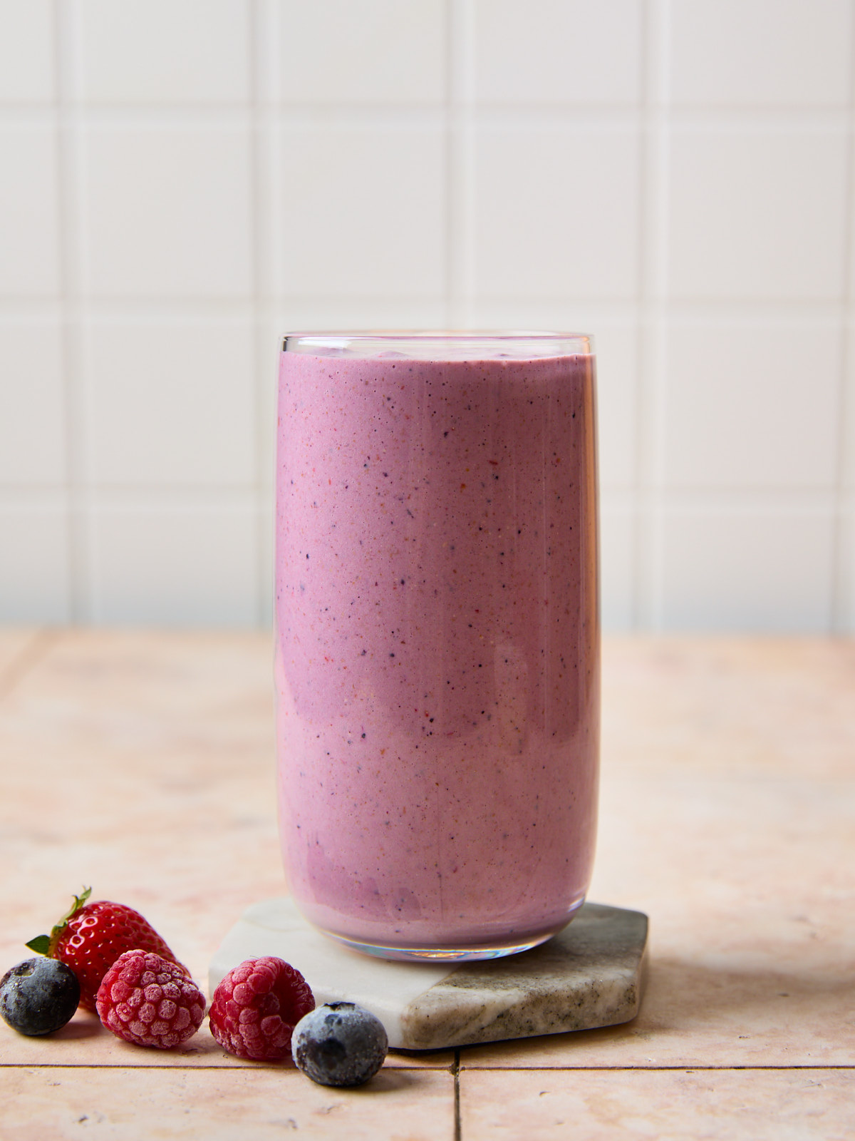 front on view of a creamy mixed berry smoothie in a glass on a marble coaster, styled with frozen berries for a fresh, vibrant look.