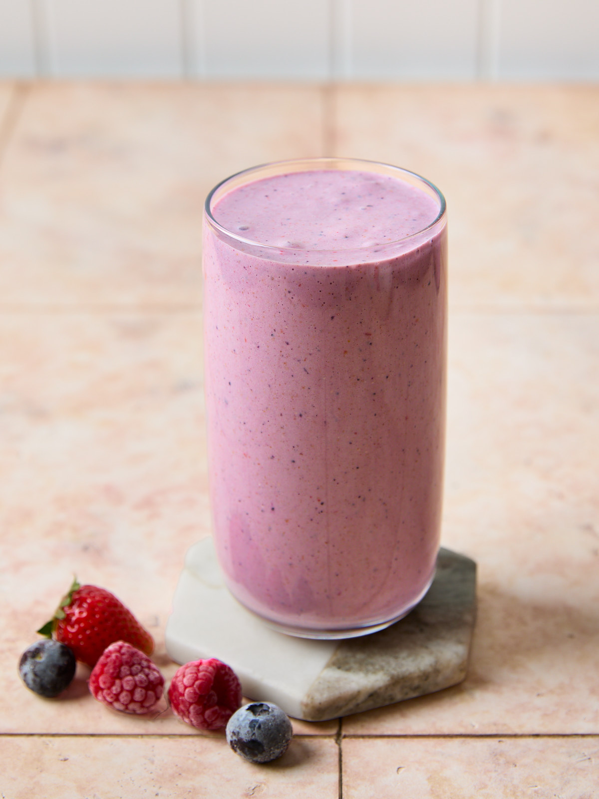 45 degree angled image of Tall glass of thick mixed berry peanut butter smoothie sitting on a marble coaster with frozen strawberries, raspberries, blueberries around it.