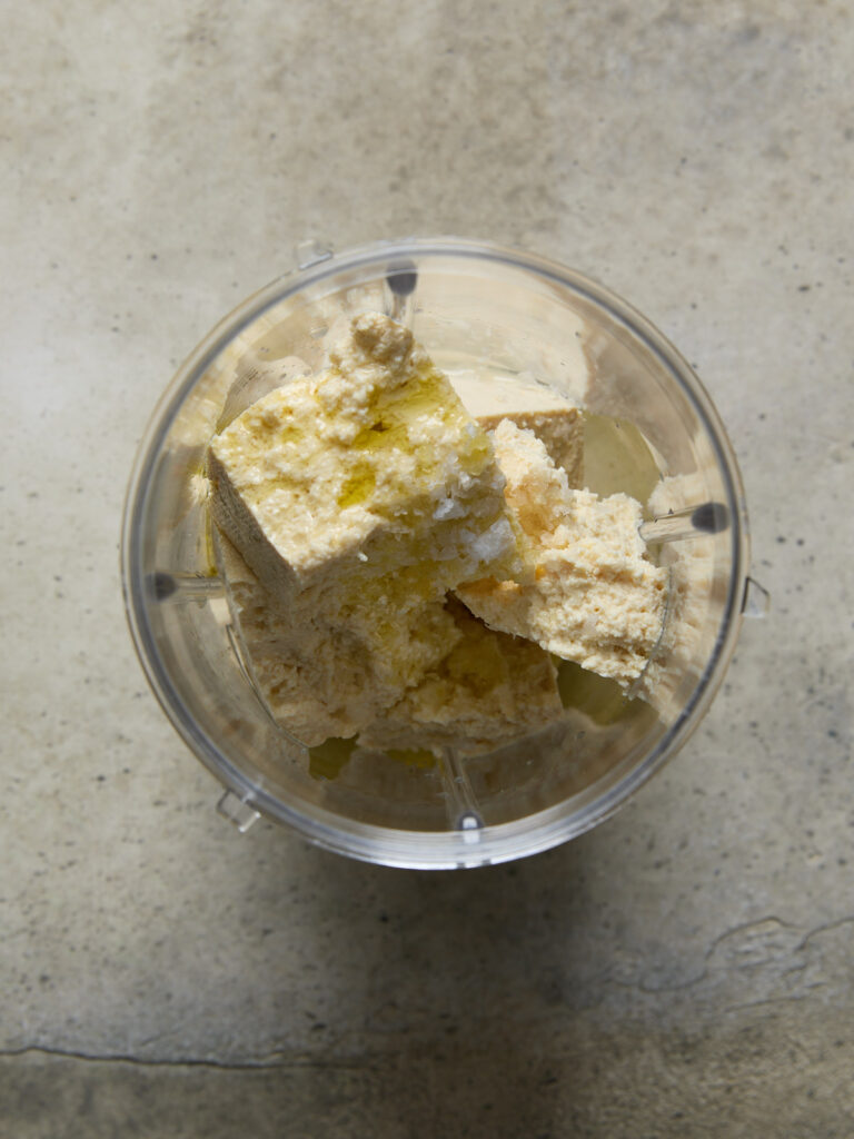 Chunks of firm tofu, olive oil, and salt in a blender cup, ready to be blended into creamy vegan ricotta.

