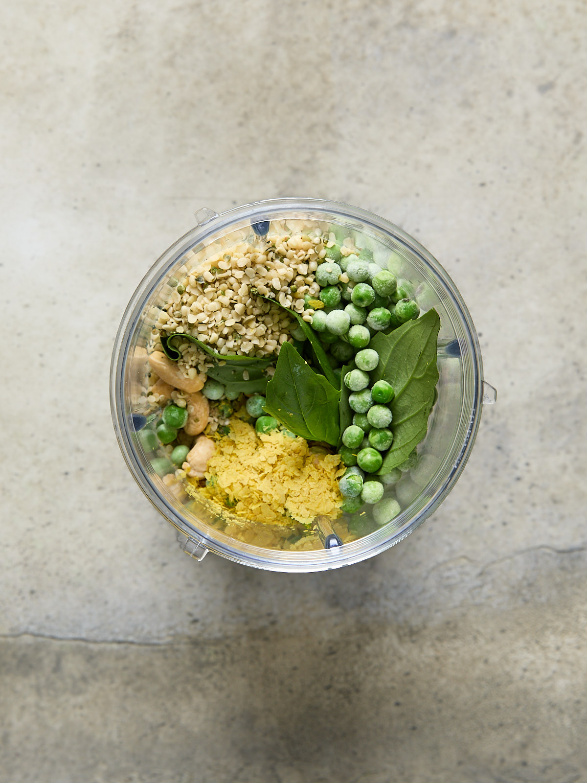 Ingredients for green pasta sauce-peas, basil, hemp seeds, nutritional yeast, and cashews-layered in a blender cup.