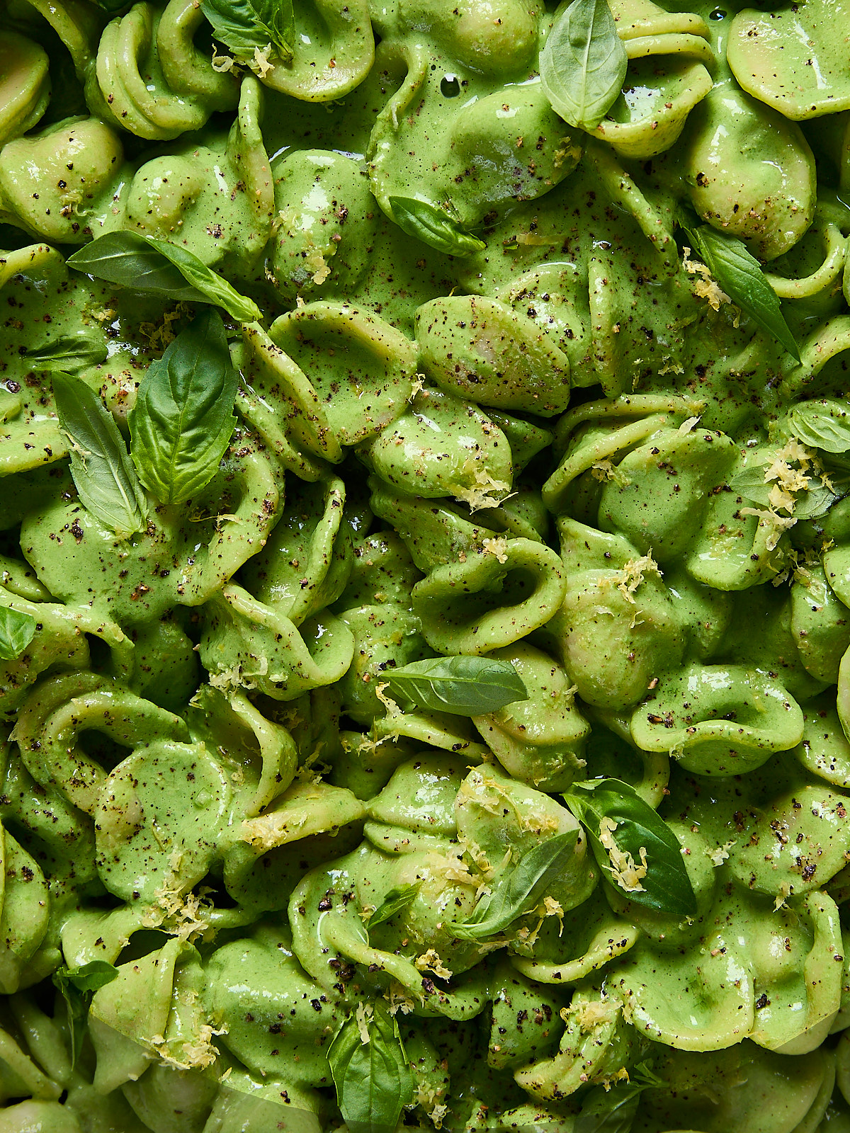 Close-up of creamy green vegan pasta with lemon zest, black pepper, and fresh basil leaves scattered on top.