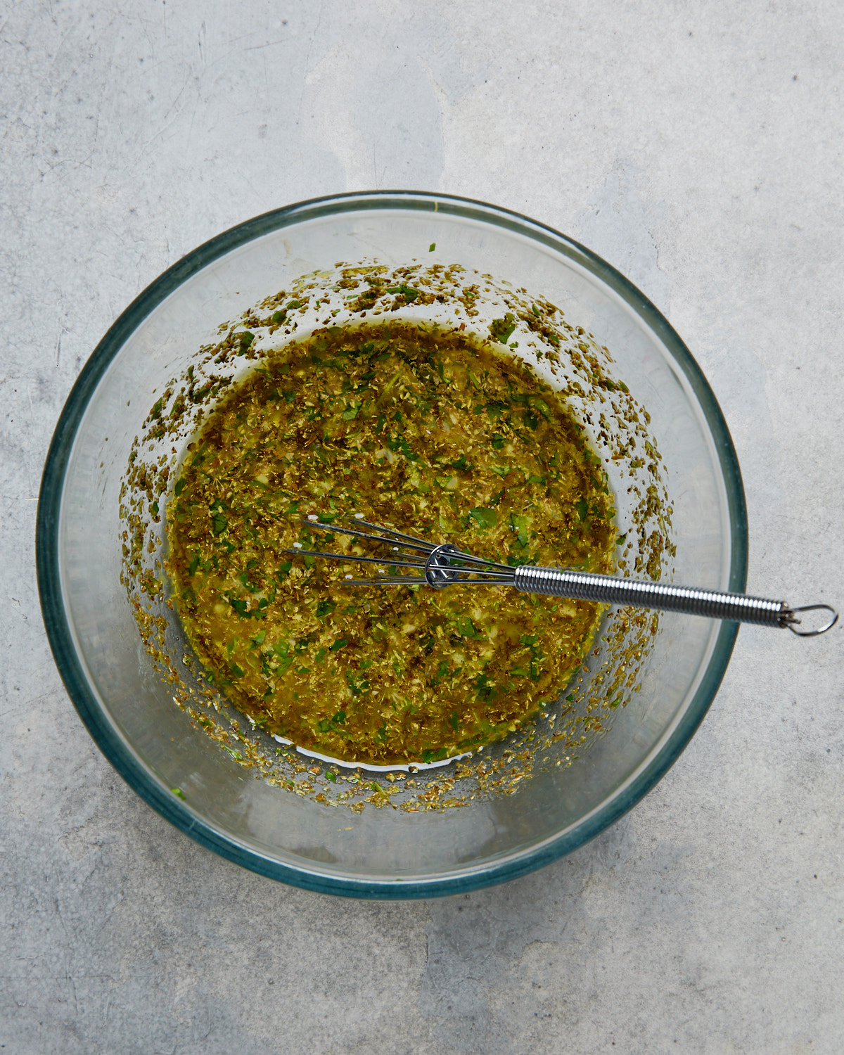 Glass mixing bowl filled with Greek marinade made from olive oil, lemon juice, herbs, and garlic, with a whisk resting inside.