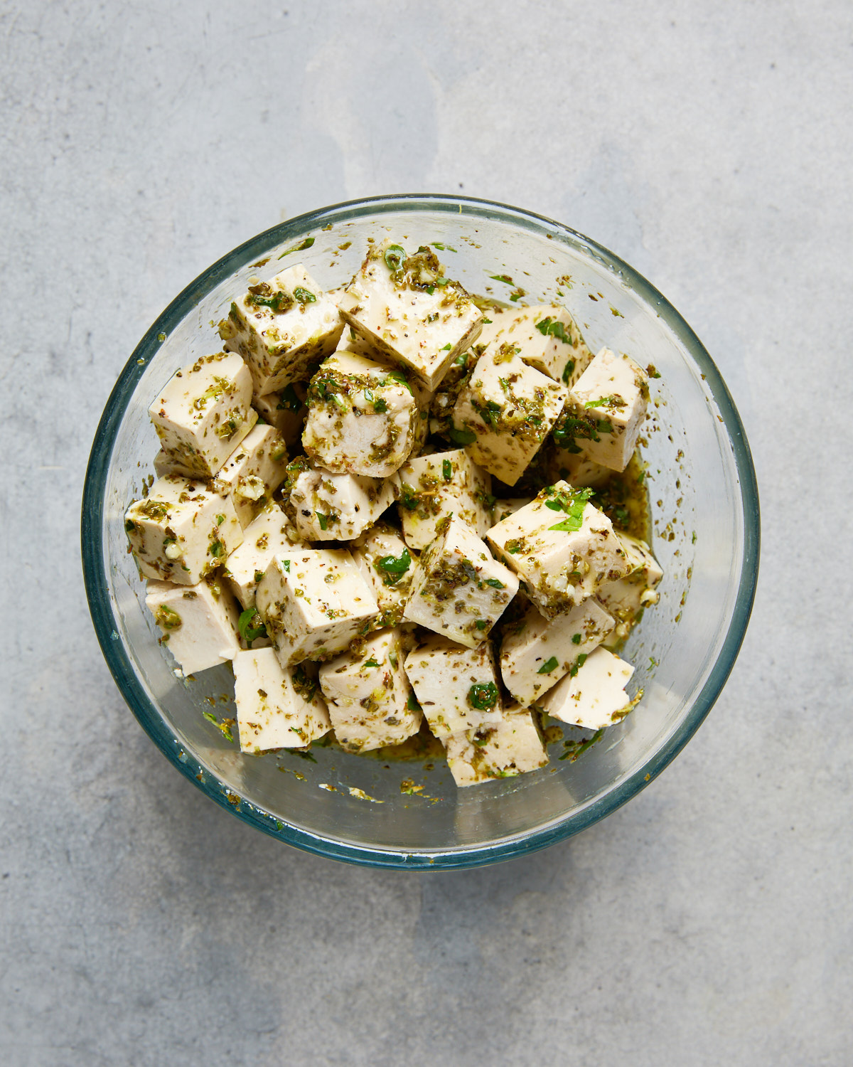 Cubed tofu coated in a herby Greek marinade inside a glass bowl, ready to be marinated.
