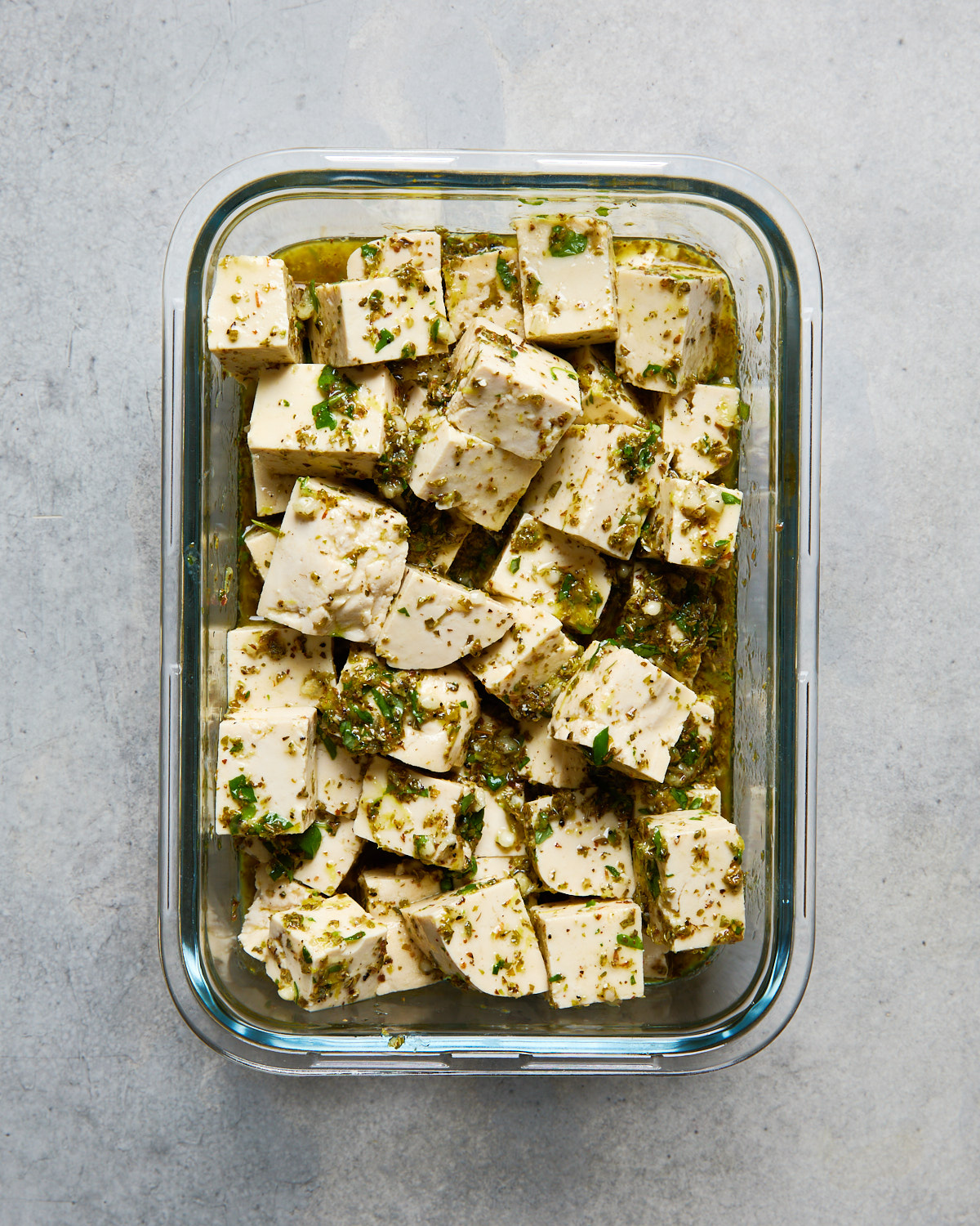 Tofu cubes marinating in olive oil, lemon, garlic, and fresh herbs in a rectangular glass container, perfect for prepping ahead.