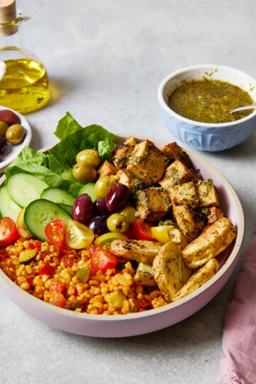 Greek-inspired grain bowl filled with bulgur wheat, cherry tomatoes, olives, cucumber, lettuce, and herby baked tofu, with a drizzle of leftover marinade on the side.