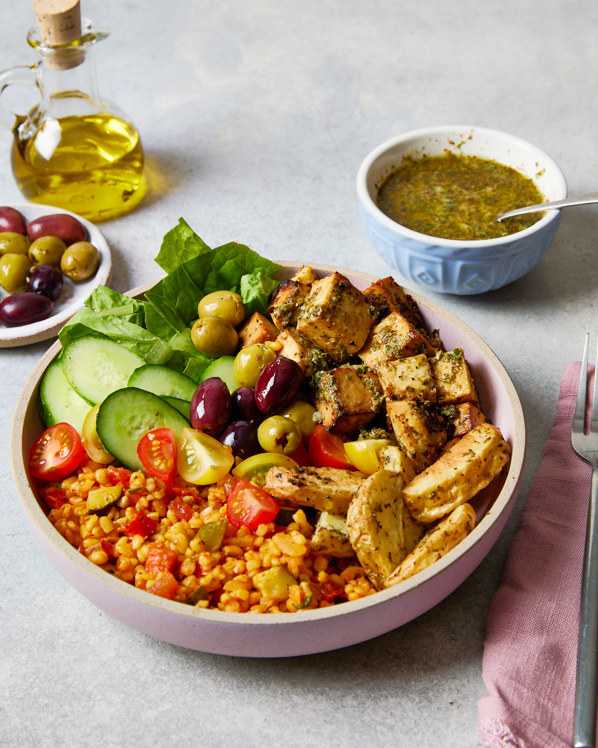 Greek-inspired grain bowl filled with bulgur wheat, cherry tomatoes, olives, cucumber, lettuce, and herby baked tofu, with a drizzle of leftover marinade on the side.