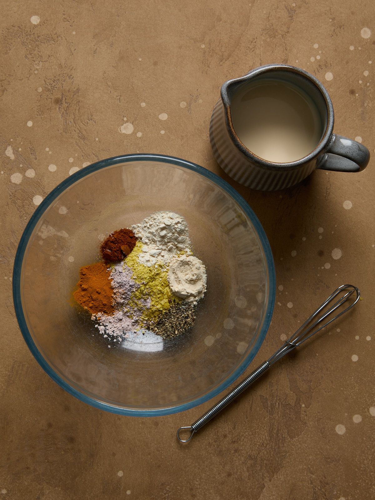 A glass mixing bowl filled with dry seasonings for vegan tofu scramble-nutritional yeast, kala namak, garlic powder, turmeric, paprika, and black pepper-next to a jug of unsweetened soy milk and a small metal whisk.