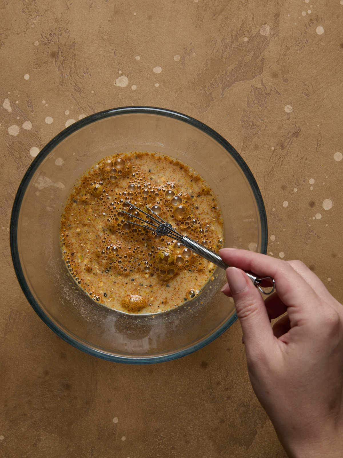 Hand whisking the tofu scramble seasoning mix with soy milk in a glass bowl, forming a creamy golden eggy sauce.