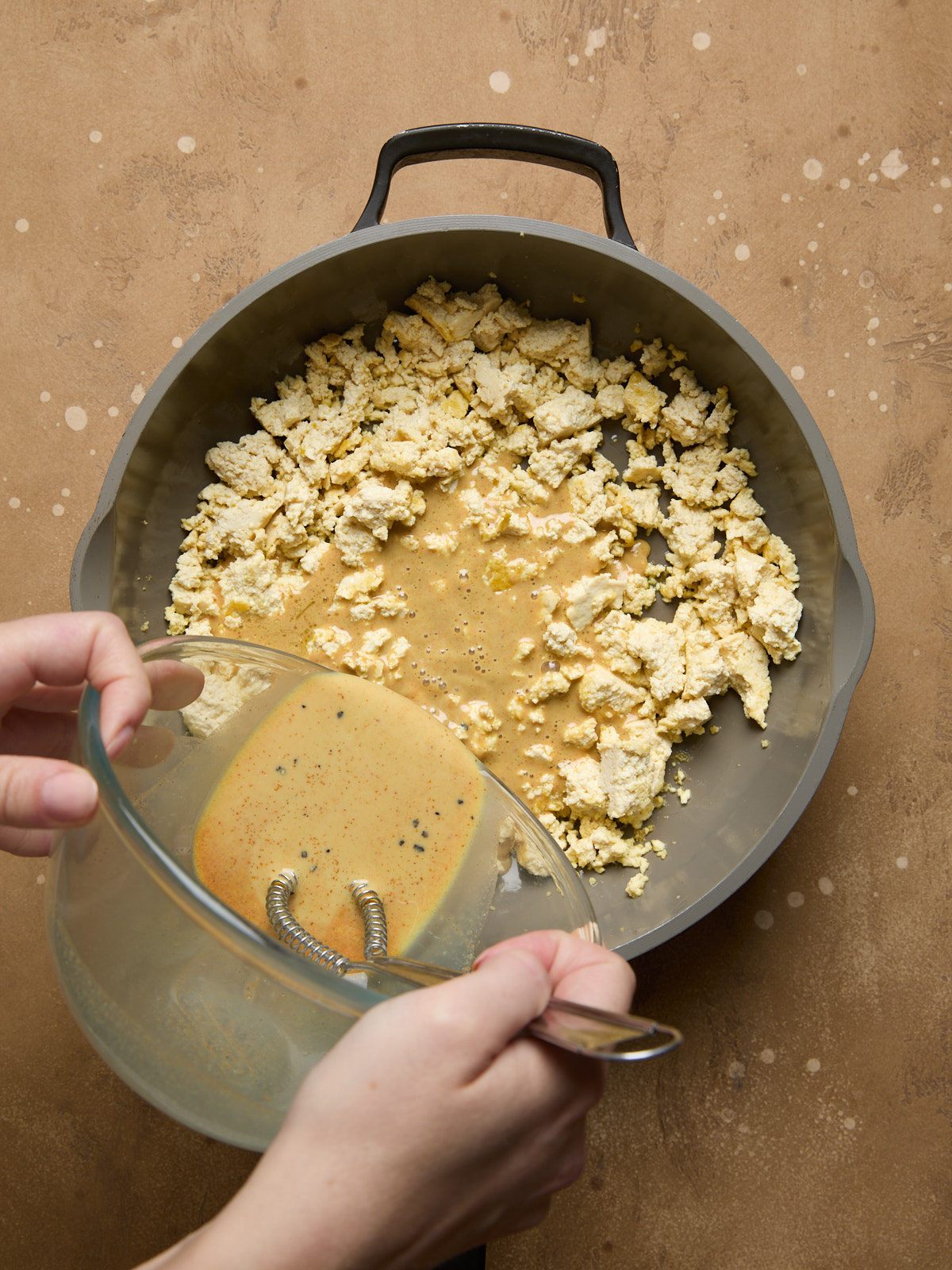 Eggy plant-based sauce being poured from a glass bowl into the pan of fried tofu to coat and flavor it.