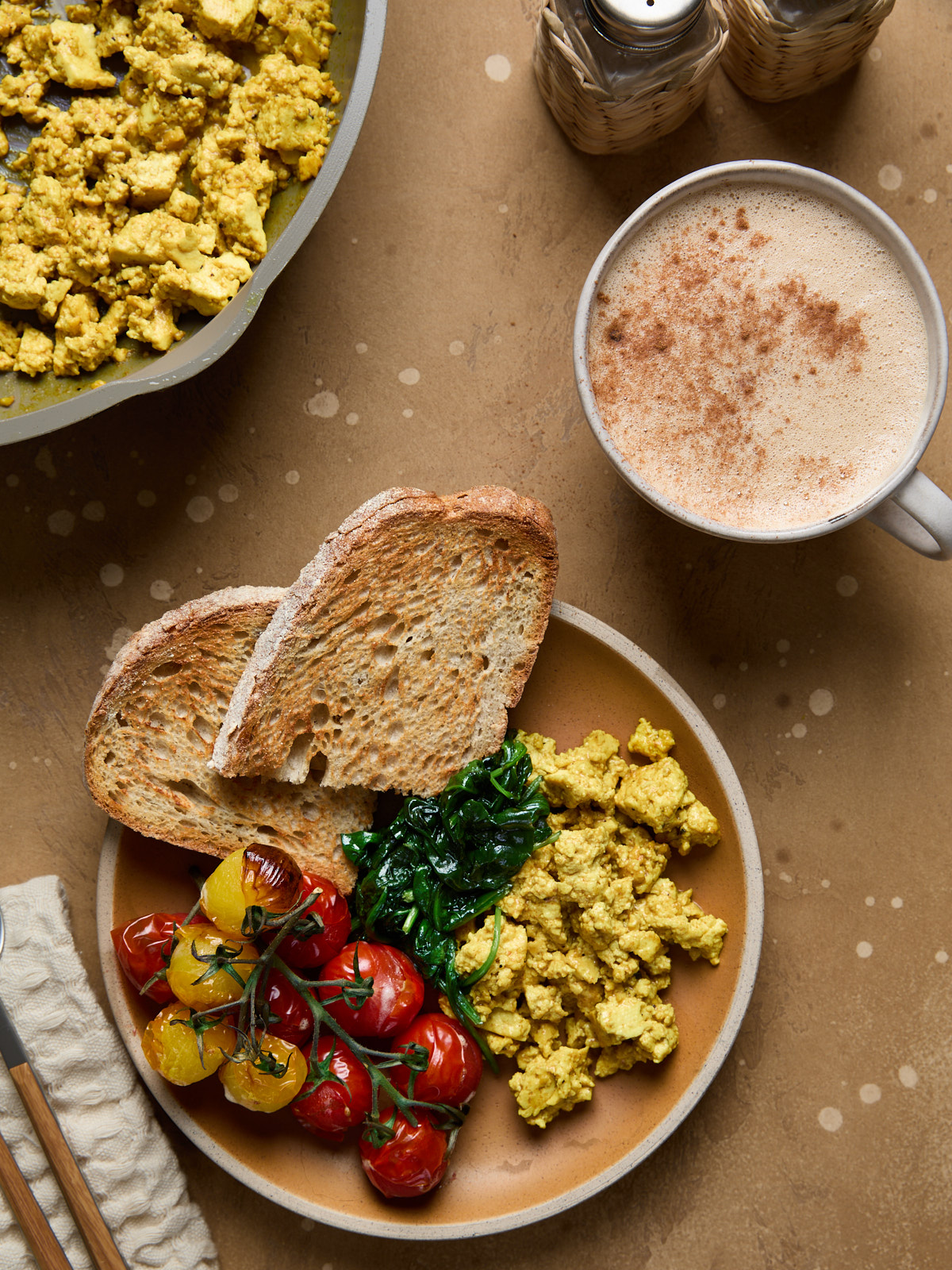 Tofu scramble served on a plate with toasted sourdough, wilted spinach, and roasted cherry tomatoes on the vine-next to a cinnamon-dusted oat milk latte.